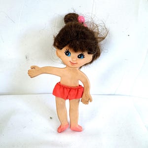 Puede incluir: Una muñeca vintage con cabello castaño recogido en un moño, adornada con una cuenta rosa. La muñeca tiene ojos azules, tez bronceada y viste un pantalón corto rojo. Los brazos de la muñeca están extendidos y sus pies son rosas.