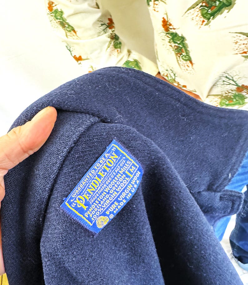 Puede incluir: Primer plano de un abrigo de lana azul marino con una etiqueta Pendleton. La etiqueta es azul y amarilla con el texto "Pendleton" y "100% Virgin Wool". El abrigo est&aacute; hecho en los EE. UU.