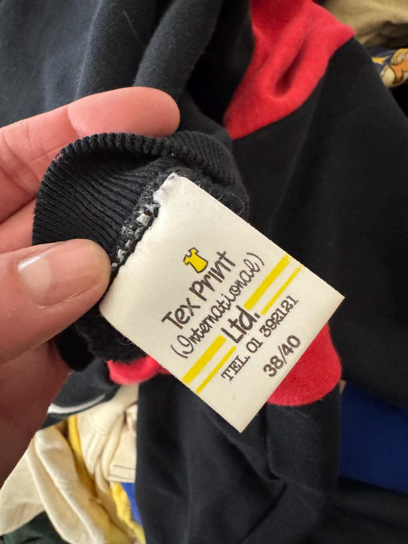 May include: Black long-sleeve shirt with red shoulder accents. The shirt has a ribbed collar and a tag that reads "Tex Print (International) Ltd." with a phone number and size 38/40.