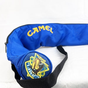 May include: A bright blue cylindrical bag featuring the yellow text CAMEL printed on the side. The circular end panel displays a colorful graphic patch with the text CAMEL and Smooth character. A black adjustable strap is attached to the bag.