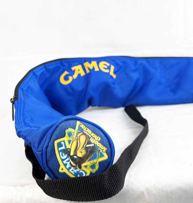 May include: A bright blue cylindrical bag featuring the yellow text CAMEL printed on the side. The circular end panel displays a colorful graphic patch with the text CAMEL and Smooth character. A black adjustable strap is attached to the bag.