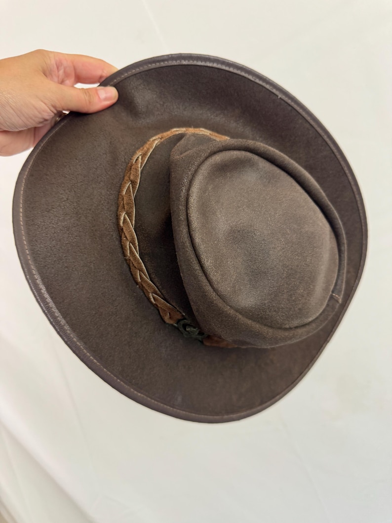 Puede incluir: Un sombrero de color marr&oacute;n oscuro con ala ancha y copa redondeada. El sombrero presenta una banda trenzada alrededor de la base de la copa, de un tono marr&oacute;n m&aacute;s claro. El material tiene un acabado mate y texturizado en toda la superficie.