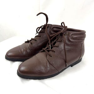May include: A pair of brown leather ankle boots with laces. The boots have a rounded toe and a low, black sole. The laces are brown and tied in a bow. The boots are on a white surface.