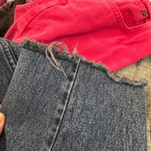 Puede incluir: Primer plano de unos vaqueros de mezclilla azul con un dobladillo deshilachado. Los vaqueros son de un azul medio con costuras visibles. Una prenda rosa brillante es visible en el fondo, junto con otras telas.