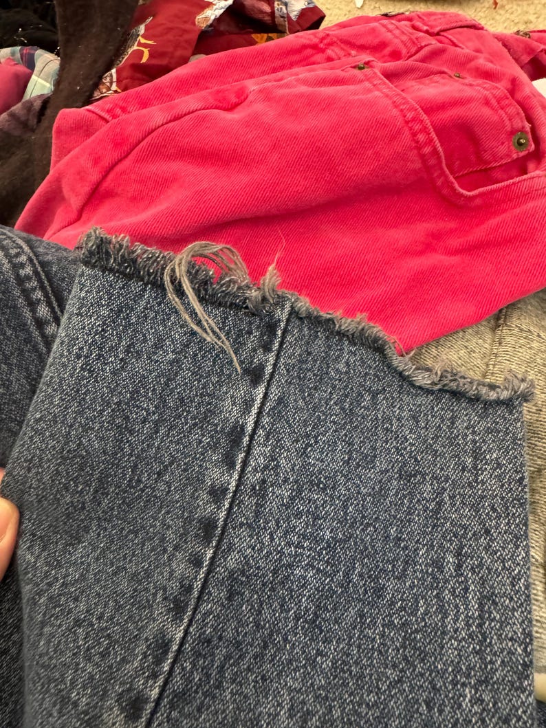 Puede incluir: Primer plano de unos vaqueros de mezclilla azul con un dobladillo deshilachado. Los vaqueros son de un azul medio con costuras visibles. Una prenda rosa brillante es visible en el fondo, junto con otras telas.
