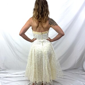 Puede incluir: Vestido sin tirantes de color marfil con un corpi&ntilde;o ajustado y una falda amplia y fluida. El vestido presenta una delicada superposici&oacute;n de encaje, un ribete dorado y un cierre de cremallera en la espalda. El vestido es hasta la rodilla y parece vintage.