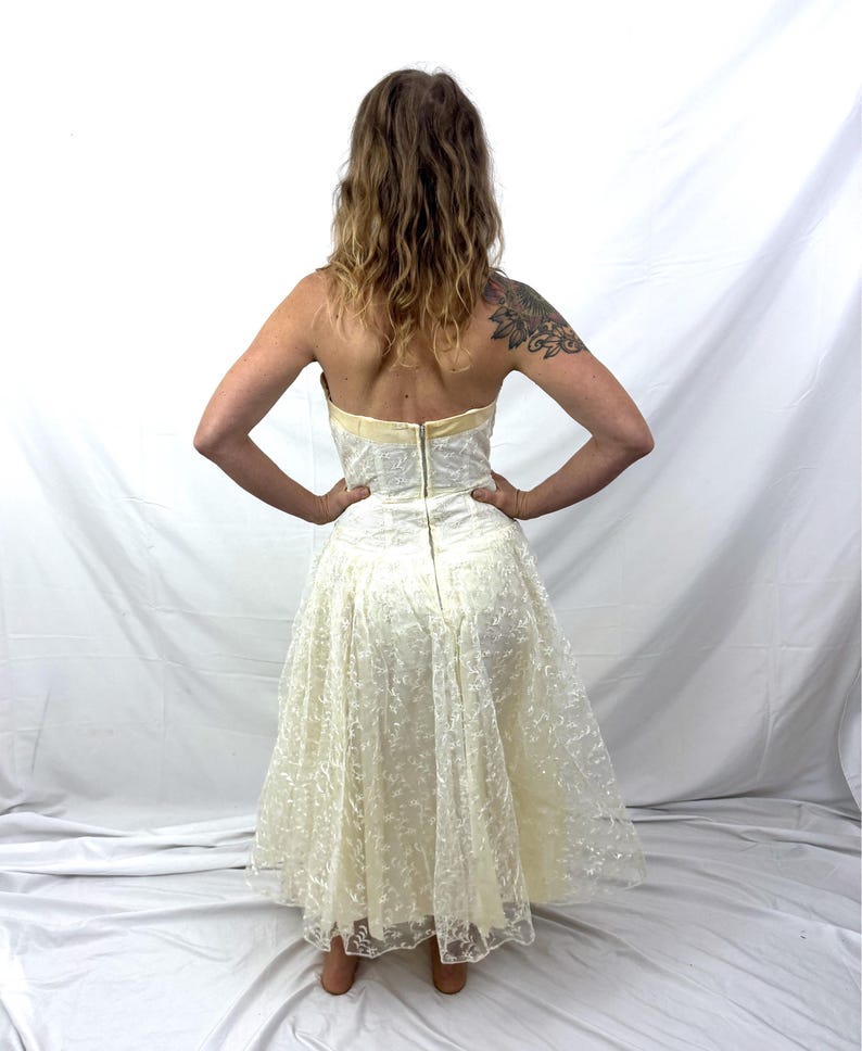 Puede incluir: Vestido sin tirantes de color marfil con un corpi&ntilde;o ajustado y una falda amplia y fluida. El vestido presenta una delicada superposici&oacute;n de encaje, un ribete dorado y un cierre de cremallera en la espalda. El vestido es hasta la rodilla y parece vintage.