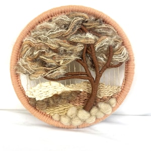 May include: A round, woven wall hanging featuring a tree with a brown trunk and branches, surrounded by textured, beige and brown fibers. The artwork is framed by a peach-colored border, creating a natural, earthy aesthetic.