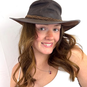 Puede incluir: Un sombrero fedora de color marr&oacute;n oscuro con ala ancha y una banda de cuero trenzado alrededor de la copa. El sombrero tiene una apariencia estructurada pero flexible con la parte superior pellizcada y un acabado mate liso en el fieltro exterior.