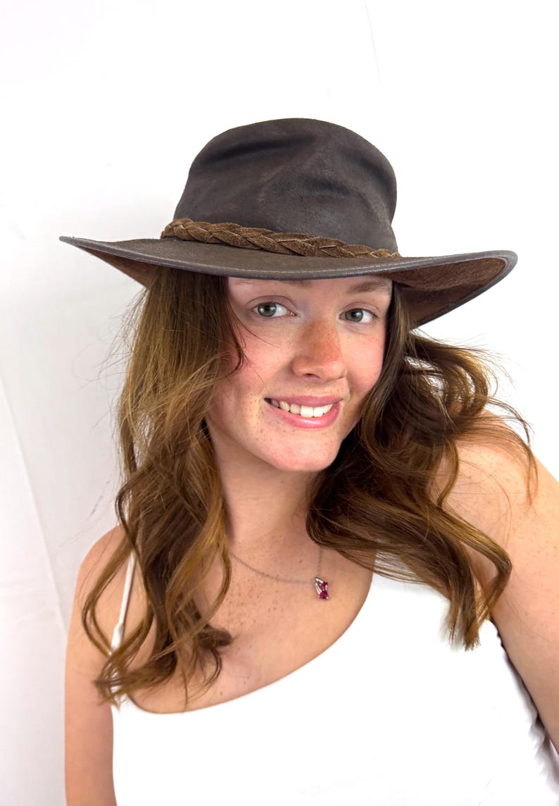 Puede incluir: Un sombrero fedora de color marr&oacute;n oscuro con ala ancha y una banda de cuero trenzado alrededor de la copa. El sombrero tiene una apariencia estructurada pero flexible con la parte superior pellizcada y un acabado mate liso en el fieltro exterior.