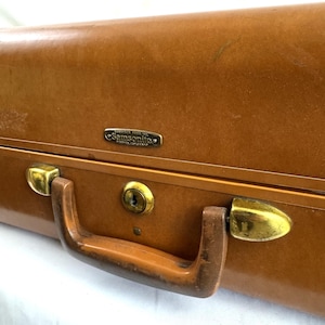 May include: A tan hardshell suitcase featuring a gold-toned metal handle, gold-toned latches, and a central key lock. A small rectangular metal plaque is attached to the front, displaying the text Samsonite and Shwayder Bros. Inc. Denver, Colorado.