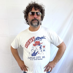 Puede incluir: Camiseta gris claro con un gr&aacute;fico que dice "Country Rock" en rojo y azul, con una mujer con sombrero de vaquero. El texto "Kansas City, MO" y un n&uacute;mero de tel&eacute;fono tambi&eacute;n est&aacute;n impresos en la camiseta.