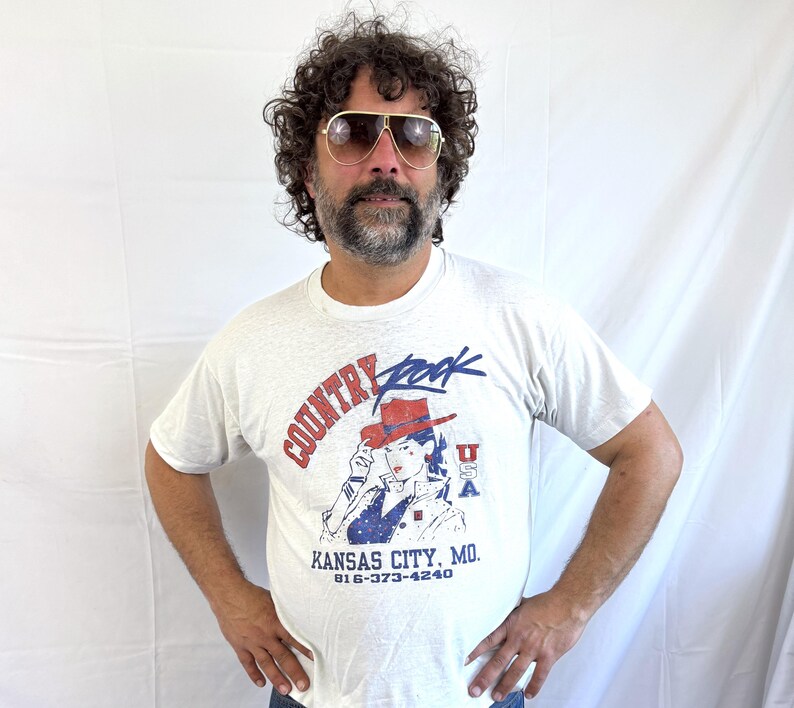 Puede incluir: Camiseta gris claro con un gr&aacute;fico que dice "Country Rock" en rojo y azul, con una mujer con sombrero de vaquero. El texto "Kansas City, MO" y un n&uacute;mero de tel&eacute;fono tambi&eacute;n est&aacute;n impresos en la camiseta.