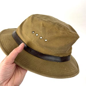 May include: A khaki-colored hat with a dark brown leather band and four ventilation eyelets. The hat has a wide brim and a slightly distressed appearance, suggesting it's made for outdoor use.