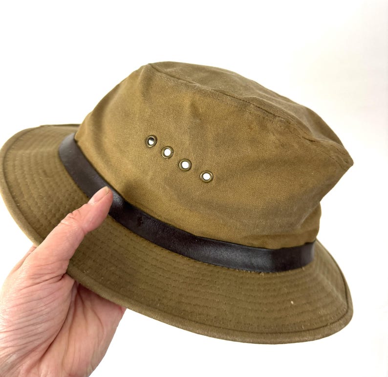 May include: A khaki-colored hat with a dark brown leather band and four ventilation eyelets. The hat has a wide brim and a slightly distressed appearance, suggesting it's made for outdoor use.