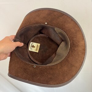 Puede incluir: Un sombrero de ante marr&oacute;n con ala ancha y forro interior de color marr&oacute;n oscuro. Una etiqueta rectangular blanca dentro de la copa presenta el texto "SQUASHY" y "GRIZZLY AUSTRALIA MADE IN TASMANIA" con un peque&ntilde;o gr&aacute;fico de un sombrero.