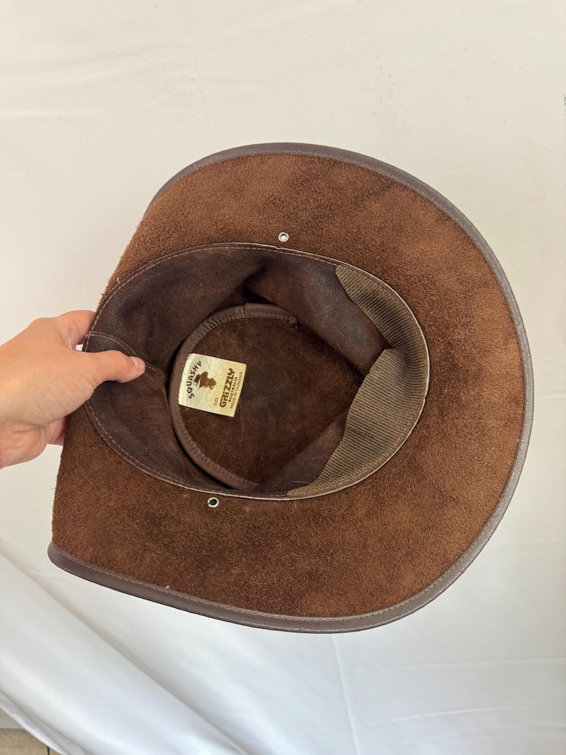 Puede incluir: Un sombrero de ante marr&oacute;n con ala ancha y forro interior de color marr&oacute;n oscuro. Una etiqueta rectangular blanca dentro de la copa presenta el texto "SQUASHY" y "GRIZZLY AUSTRALIA MADE IN TASMANIA" con un peque&ntilde;o gr&aacute;fico de un sombrero.