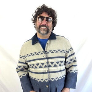 May include: A beige and navy blue patterned jacket with a dark blue collar and buttons. The jacket features a geometric design with horizontal stripes and triangle shapes. The jacket is worn over a blue shirt.