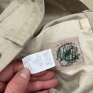 Puede incluir: Un pantal&oacute;n beige Filson con un parche de cuero marr&oacute;n que dice "A Genuine Filson" y una etiqueta blanca que dice "C.C. Filson Co. Seattle, WA Made in USA Style 575 2001/04 3749 Size 30 100% cotton Care on reverse"