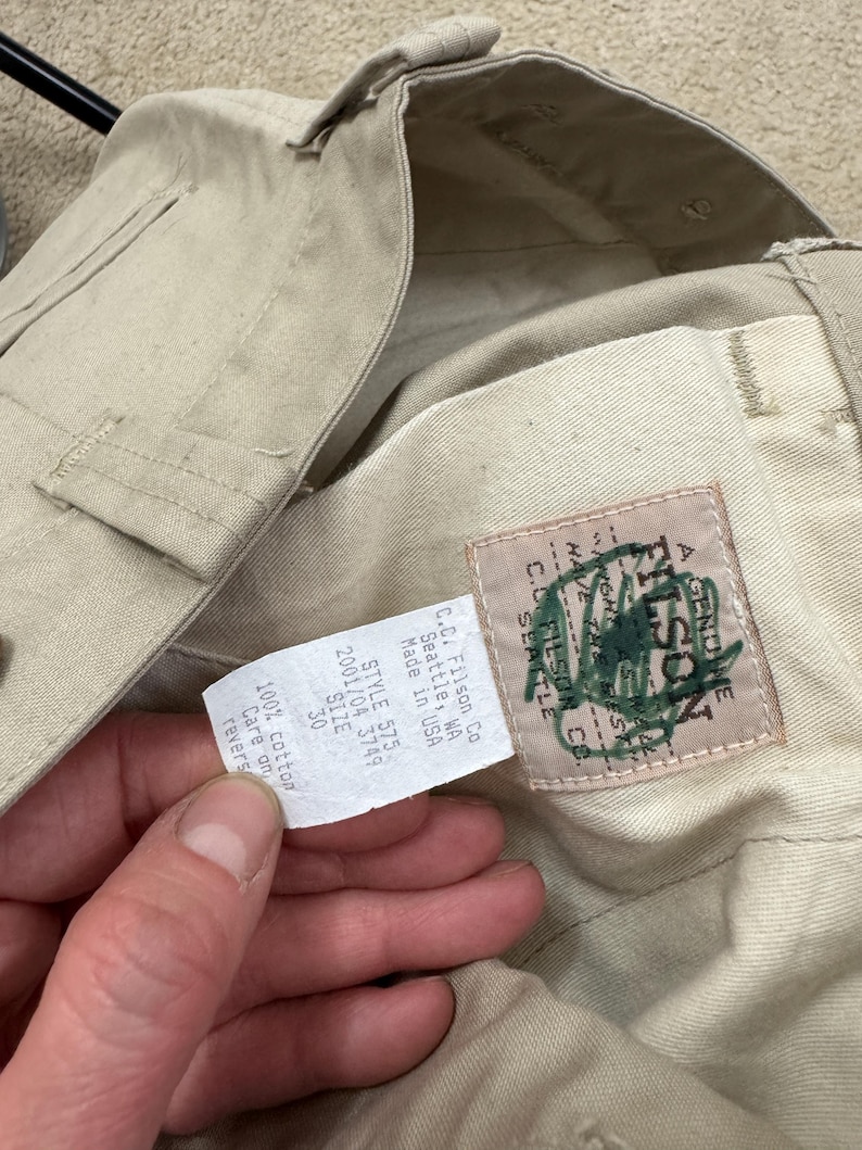 Puede incluir: Un pantal&oacute;n beige Filson con un parche de cuero marr&oacute;n que dice "A Genuine Filson" y una etiqueta blanca que dice "C.C. Filson Co. Seattle, WA Made in USA Style 575 2001/04 3749 Size 30 100% cotton Care on reverse"