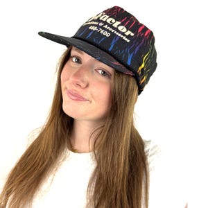 May include: A black trucker hat with a rainbow-colored design and the word "Factor" in white. The hat also features the text "Fashions & Accessories 488-7600". The hat has a black brim and a black rope detail.