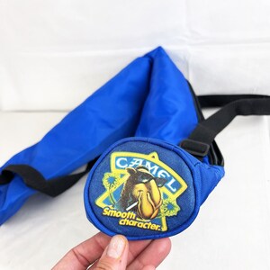 May include: A bright blue nylon sling bag featuring a circular patch on the front. The patch displays a yellow star outline, a cartoon camel wearing sunglasses, and the text CAMEL Smooth character in yellow and white lettering.