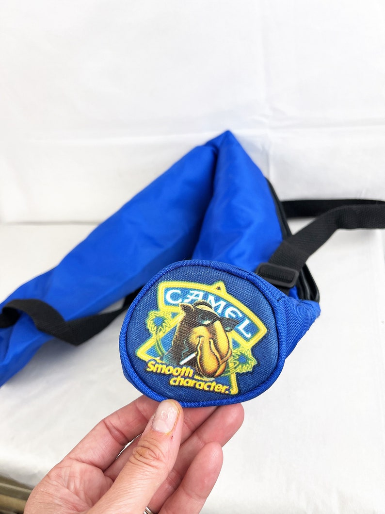 May include: A bright blue nylon sling bag featuring a circular patch on the front. The patch displays a yellow star outline, a cartoon camel wearing sunglasses, and the text CAMEL Smooth character in yellow and white lettering.