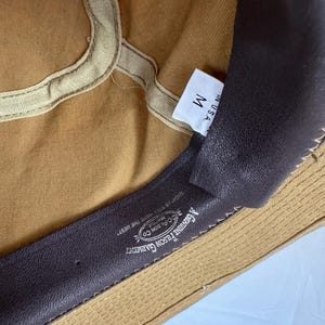 May include: Close-up of the inside of a tan canvas hat. The hat has a brown leather sweatband with the text "A GENUINE FILSON GARMENT" and "FILSON CO." The tag reads "M" and "IN U.S.A."