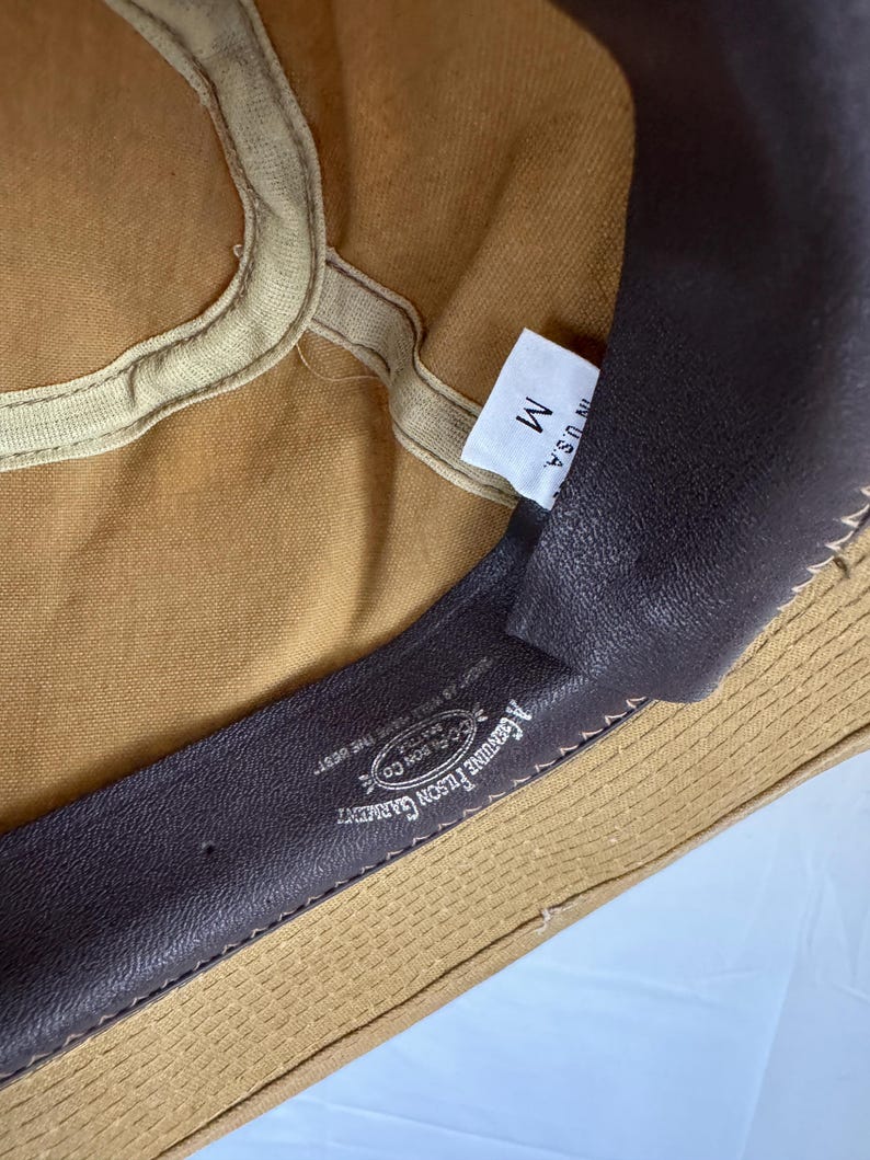 May include: Close-up of the inside of a tan canvas hat. The hat has a brown leather sweatband with the text "A GENUINE FILSON GARMENT" and "FILSON CO." The tag reads "M" and "IN U.S.A."