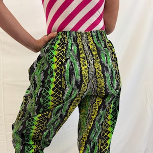 May include: Black pants with a vibrant green and yellow geometric pattern. The pants have an elastic waistband and a loose fit. The top is a pink and white striped tank top. The overall style is retro and eye-catching.