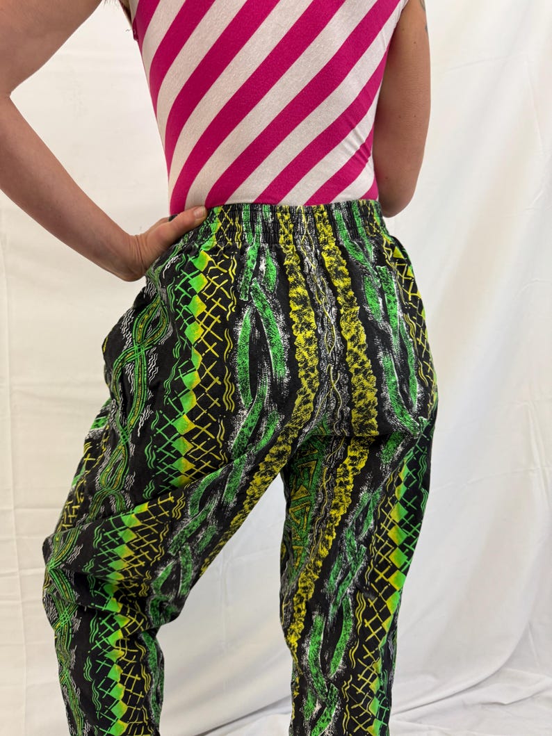 May include: Black pants with a vibrant green and yellow geometric pattern. The pants have an elastic waistband and a loose fit. The top is a pink and white striped tank top. The overall style is retro and eye-catching.