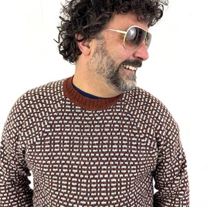 Puede incluir: Un su&eacute;ter estampado en marr&oacute;n, blanco y rojo oscuro. El su&eacute;ter tiene cuello redondo y un dise&ntilde;o geom&eacute;trico. La persona lleva gafas de sol de aviador con montura dorada. El fondo es blanco liso.