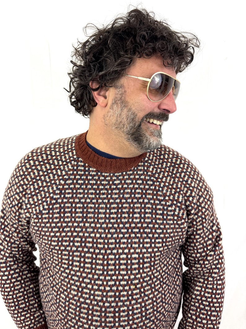 Puede incluir: Un su&eacute;ter estampado en marr&oacute;n, blanco y rojo oscuro. El su&eacute;ter tiene cuello redondo y un dise&ntilde;o geom&eacute;trico. La persona lleva gafas de sol de aviador con montura dorada. El fondo es blanco liso.