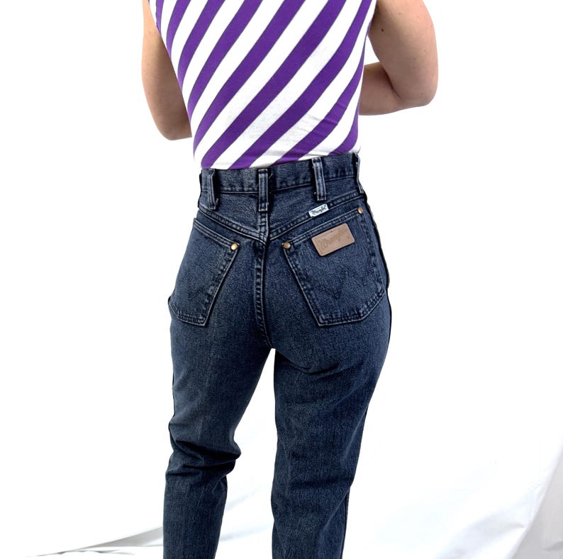 Puede incluir: Vaqueros de mezclilla de lavado oscuro con cintura alta y etiqueta Wrangler. Los vaqueros tienen un dise&ntilde;o cl&aacute;sico de cinco bolsillos y remaches de color lat&oacute;n. Tambi&eacute;n se ve una camiseta sin mangas con rayas diagonales blancas y moradas.