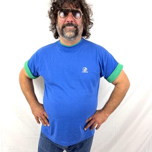 Puede incluir: Una camiseta azul con ribetes verdes en el cuello y las mangas. La camiseta tiene un peque&ntilde;o logotipo en el pecho. La persona de la imagen lleva vaqueros azules y gafas de sol de aviador.