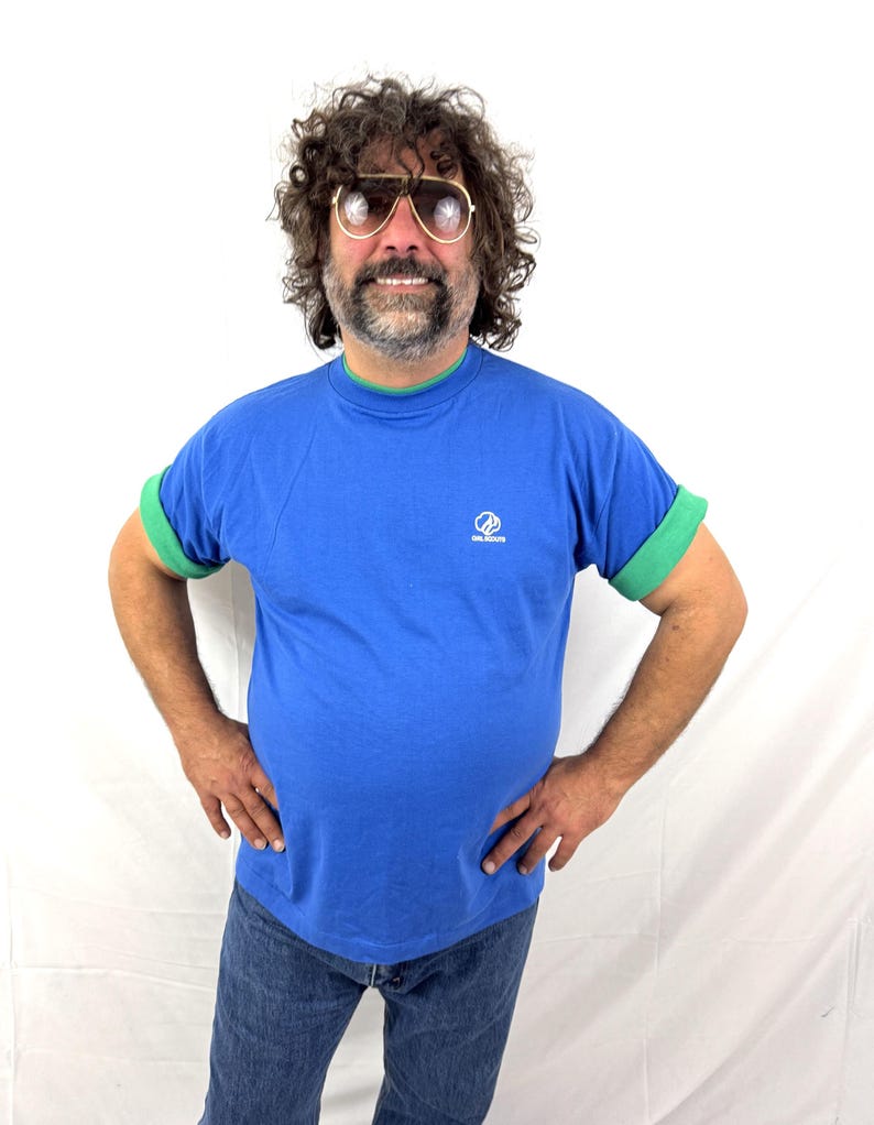 Puede incluir: Una camiseta azul con ribetes verdes en el cuello y las mangas. La camiseta tiene un peque&ntilde;o logotipo en el pecho. La persona de la imagen lleva vaqueros azules y gafas de sol de aviador.