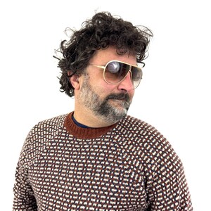 Puede incluir: Gafas de sol de aviador con montura dorada y lentes degradadas marrones. Las gafas de sol se llevan con un su&eacute;ter estampado en marr&oacute;n, blanco y azul marino. El su&eacute;ter tiene cuello redondo marr&oacute;n y una camiseta interior azul marino.