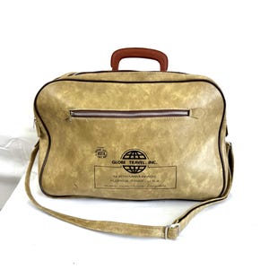 May include: A tan leather travel bag with a brown handle and a shoulder strap. The bag has a zipper closure and a logo that reads "Globe Travel, Inc. North Miami Beach, Florida 33162 U.S.A. Cruises, Tours, Tickets, Everything".