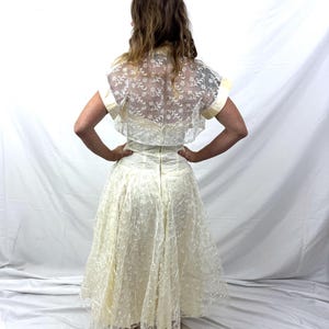 Puede incluir: Vestido vintage de color marfil con una superposici&oacute;n de encaje. El vestido presenta un corpi&ntilde;o ajustado y una falda amplia. Un bolero transparente de manga corta con detalles de encaje floral completa el conjunto. Un vestido de estilo cl&aacute;sico.
