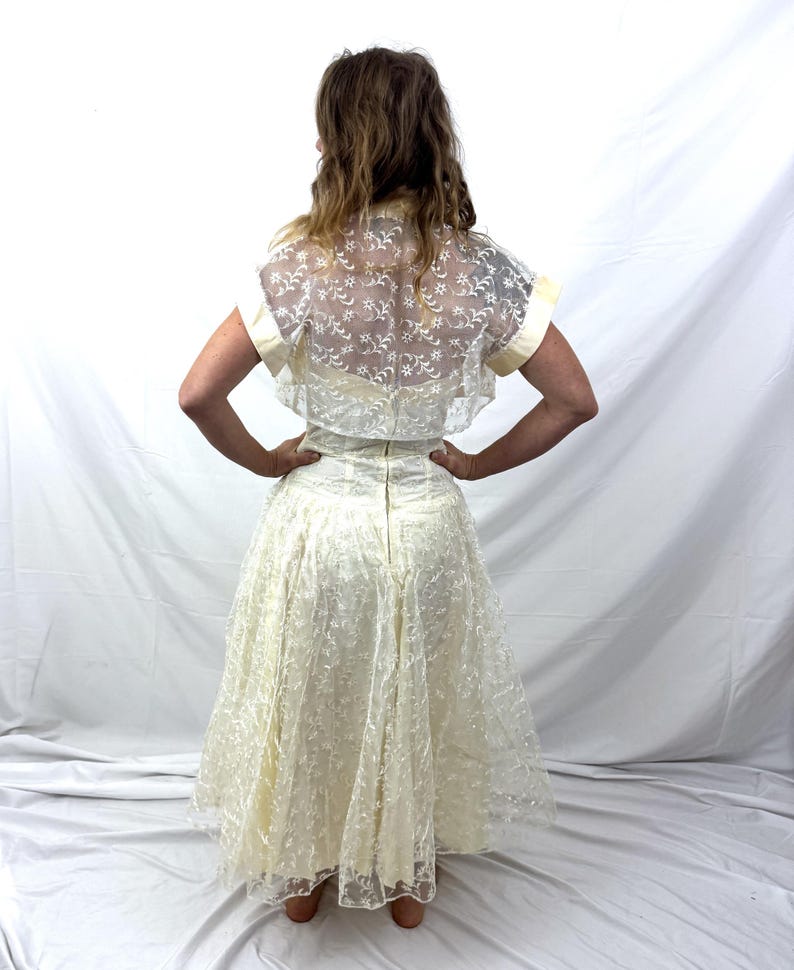 Puede incluir: Vestido vintage de color marfil con una superposici&oacute;n de encaje. El vestido presenta un corpi&ntilde;o ajustado y una falda amplia. Un bolero transparente de manga corta con detalles de encaje floral completa el conjunto. Un vestido de estilo cl&aacute;sico.