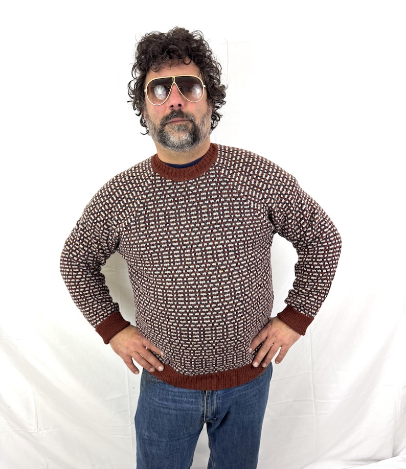 Puede incluir: Un su&eacute;ter con estampado marr&oacute;n y blanco con cuello redondo y mangas largas. El su&eacute;ter tiene pu&ntilde;os marrones y un dobladillo marr&oacute;n. La persona de la imagen lleva vaqueros azules y gafas de sol de aviador.