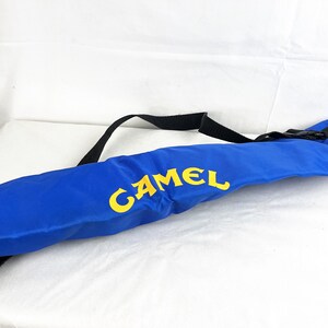 May include: A long, cylindrical blue carrying case featuring the word CAMEL printed in bold yellow letters. The bag includes a black adjustable nylon strap attached to the top, designed for carrying the item over a shoulder or across the body.