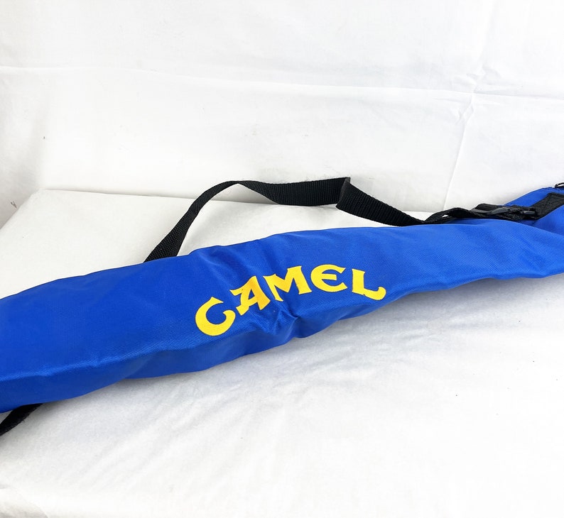 May include: A long, cylindrical blue carrying case featuring the word CAMEL printed in bold yellow letters. The bag includes a black adjustable nylon strap attached to the top, designed for carrying the item over a shoulder or across the body.