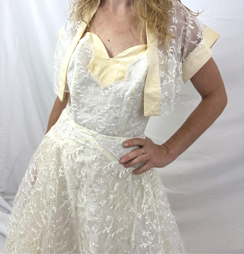 Puede incluir: Vestido de encaje marfil con escote coraz&oacute;n y bolero a juego. El vestido presenta un corpi&ntilde;o entallado y una falda acampanada. El bolero tiene mangas cortas y ribete de sat&eacute;n. Conjunto de estilo vintage.