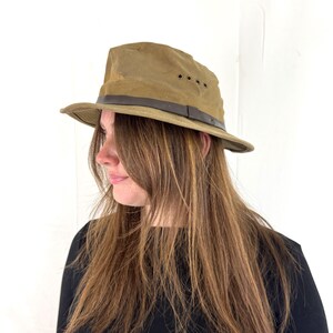 May include: A khaki-colored fedora hat with a brown leather band. The hat has a slightly distressed appearance and features three small ventilation holes on the side. The brim is curved, and the hat appears to be made of a durable, possibly waxed, material.