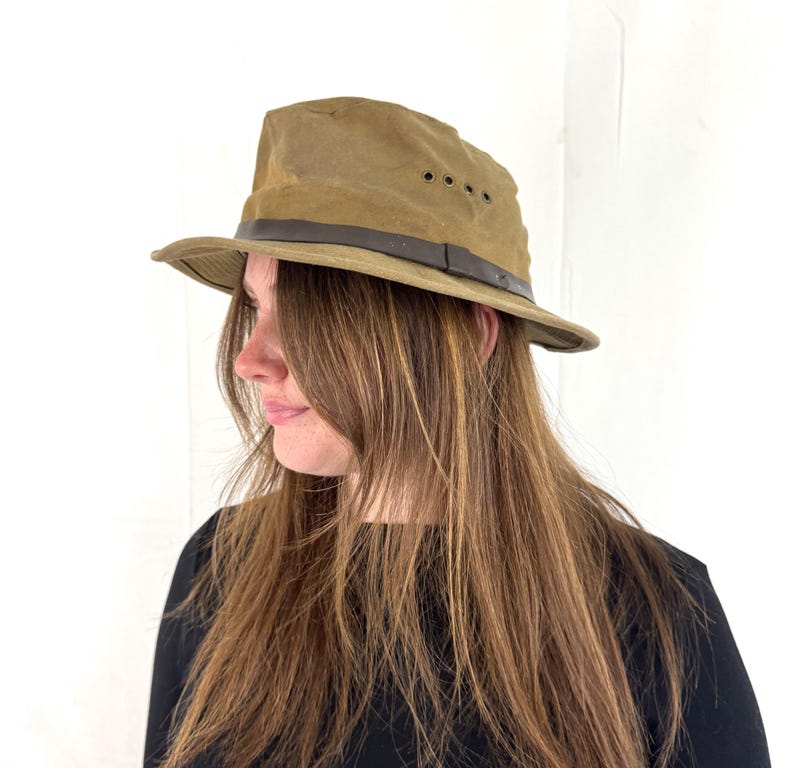 May include: A khaki-colored fedora hat with a brown leather band. The hat has a slightly distressed appearance and features three small ventilation holes on the side. The brim is curved, and the hat appears to be made of a durable, possibly waxed, material.