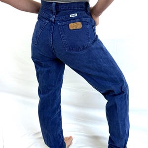 K&ouml;nnte beinhalten: Eine blaue Jeans mit hoher Taille und einem braunen Leder-Wrangler-Patch auf der Ges&auml;&szlig;tasche.