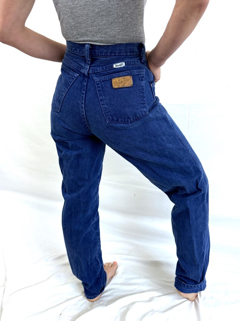 K&ouml;nnte beinhalten: Eine blaue Jeans mit hoher Taille und einem braunen Leder-Wrangler-Patch auf der Ges&auml;&szlig;tasche.