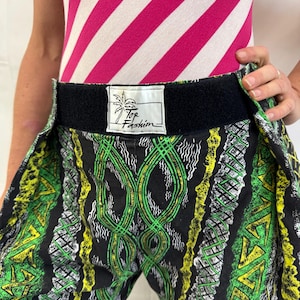 May include: Black, green, and yellow patterned pants with a black waistband. The pants feature a repeating design of geometric shapes and lines. The label on the waistband reads "Top Fashion". The top is a pink and white striped bodysuit.