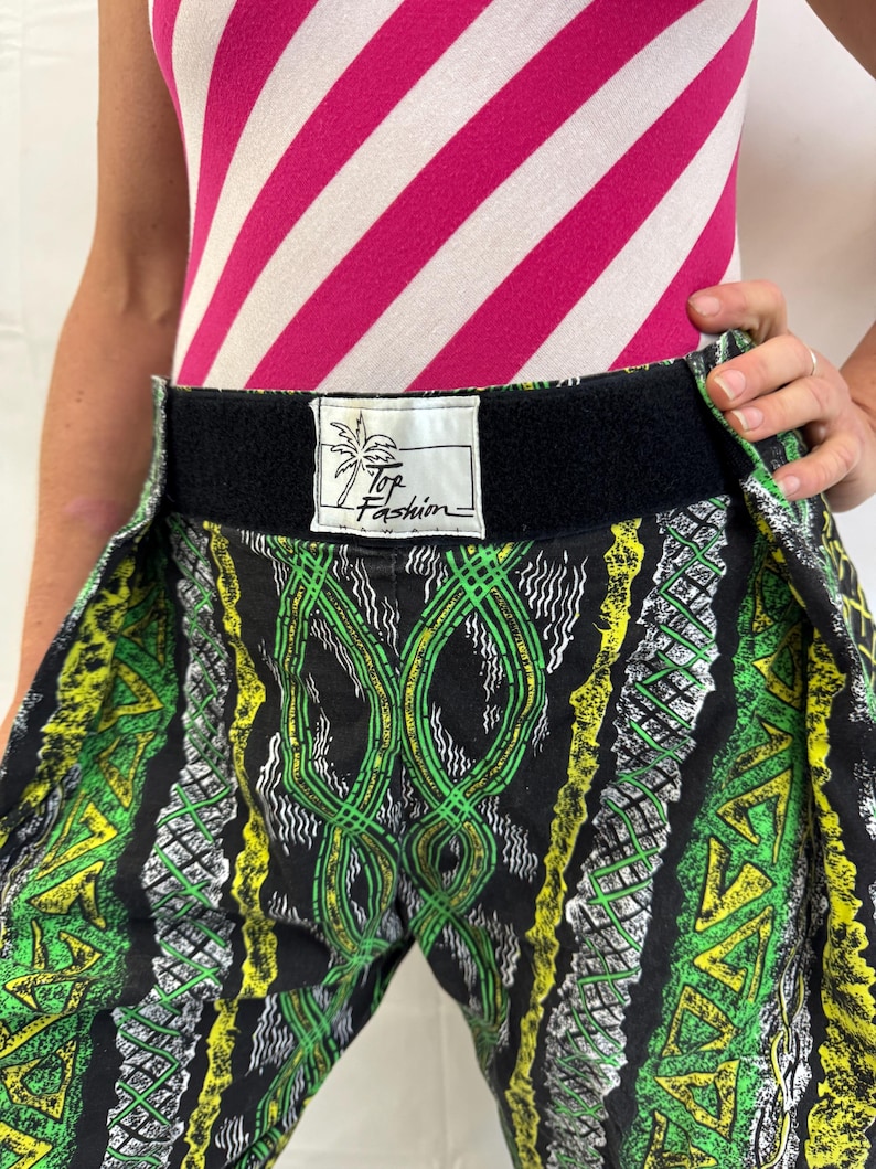 May include: Black, green, and yellow patterned pants with a black waistband. The pants feature a repeating design of geometric shapes and lines. The label on the waistband reads "Top Fashion". The top is a pink and white striped bodysuit.